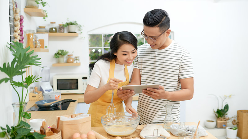 Young asian man and woman together cooking cake and bread with egg, looking menu from tablet in the flour happy relaxing in at home