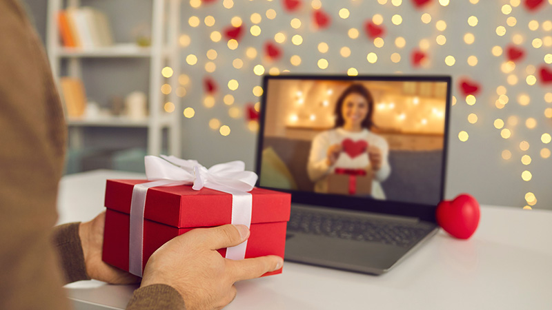 Video call on Saint Valentine's Day. Couple sending each other love and showing presents on virtual date. Closeup of man's hands holding gift box. Shimmering yellow lights, soft focus, selective focus