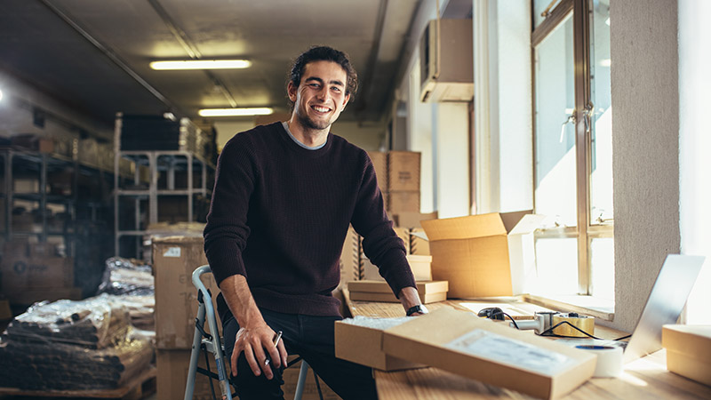 Successful online business owner sitting at his workdesk. Young man with laptop and shipment box on desk, looking at camera and smiling.
