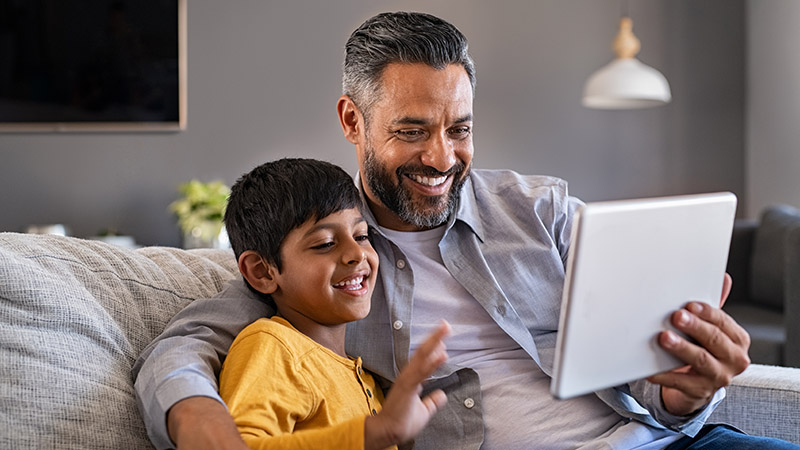 Indian father and smiling son sitting on couch using digital tablet at home. Man and boy using tablet for video calling at home. Middle eastern dad with son doing videocall during quarantine.